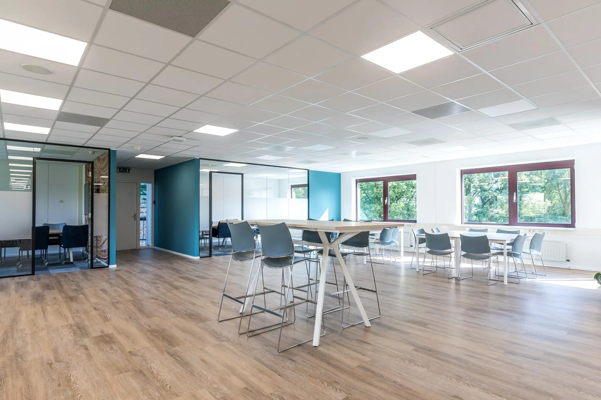 Moderne, lichte kantoorkantine aan de Baarnsche Dijk met houten vloer, blauwe accenten en grote ramen met uitzicht op groen.